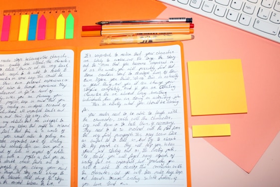 A workspace with an open notebook filled with handwritten notes. Around the notebook, there are colorful sticky notes, a ruler, several pens, and a section of a keyboard visible. The background is a warm orange and pink color scheme.