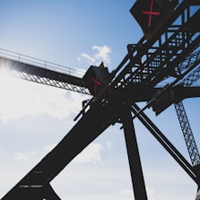 Silhouetted structure of a bridge or framework with diagonal and vertical beams against a bright blue sky. Illuminated signs with red 'X' symbols are visible on the structure, indicating some form of restriction or warning.