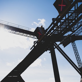 Silhouetted structure of a bridge or framework with diagonal and vertical beams against a bright blue sky. Illuminated signs with red 'X' symbols are visible on the structure, indicating some form of restriction or warning.