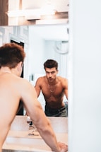 A shirtless man is looking at himself in the bathroom mirror, leaning forward with both hands on the sink. The bathroom is brightly lit with a neutral color palette, featuring a reflective wall mirror and modern fixtures. The focus is on his serious expression and toned physique.