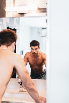 A confident man standing in a bright, modern bathroom, looking at himself in the mirror with a determined expression.