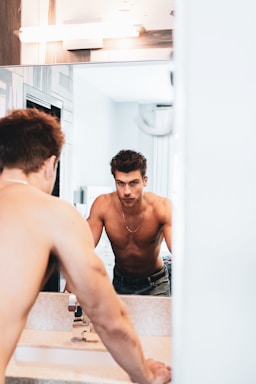 A shirtless man is looking at himself in the bathroom mirror, leaning forward with both hands on the sink. The bathroom is brightly lit with a neutral color palette, featuring a reflective wall mirror and modern fixtures. The focus is on his serious expression and toned physique.