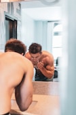 A person with short hair and a muscular build is standing shirtless at a bathroom sink, splashing water on their face. The bathroom has light-colored walls and a window with curtains. The reflection in the mirror captures the person's focused expression as water droplets scatter.