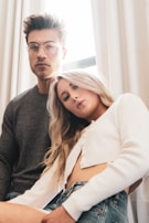A man and a woman are seated indoors with a window behind them. The man is wearing glasses and a dark sweater, and has a composed expression. The woman is wearing a white cropped top and denim shorts, leaning slightly towards the camera with a relaxed demeanor. Light filters gently through the curtains, creating a soft ambiance.