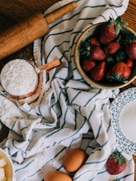 A rustic kitchen setup with a wooden rolling pin, a bowl filled with fresh strawberries, a cup of flour, eggs, and a bowl of sugar placed on a striped kitchen towel. The items are arranged in a cozy and inviting manner, suggesting baking preparations.