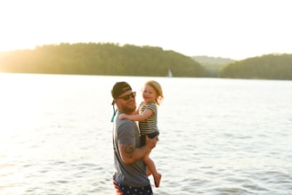 A man in a cap and sunglasses is holding a young child by the water's edge. The man is smiling, and the child appears happy, with a background of a scenic lake and trees under bright sunlight.