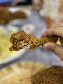 A hand holds a piece of fried chicken in focus, with a crispy, golden-brown texture. In the background, other blurred food items and a table setting can be seen.