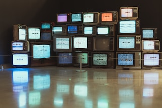 A collection of vintage television sets are stacked upon each other, each displaying static or random color bandwidth. These TVs create an artistic installation with reflections visible on the polished floor, presenting a nostalgic and retro aesthetic.