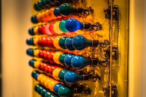 An array of vibrant light bulbs in various colors is mounted on a structure. The bulbs are arranged in three rows, each consisting of bulbs with a glossy surface, creating a striking visual contrast against the background. The wires and sockets are visible, adding a sense of industrial design.