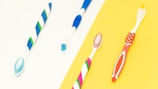 Four toothbrushes with different colorful designs are laid out against a split background of white and yellow. The toothbrushes feature combinations of blue, green, red, and pink colors.