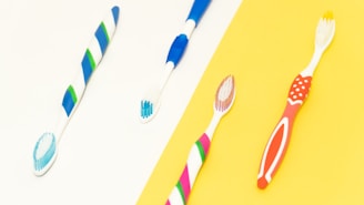 Four toothbrushes with different colorful designs are laid out against a split background of white and yellow. The toothbrushes feature combinations of blue, green, red, and pink colors.