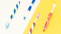 Four toothbrushes with different colorful designs are laid out against a split background of white and yellow. The toothbrushes feature combinations of blue, green, red, and pink colors.