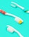 Three toothbrushes are placed on a bright turquoise background. The toothbrushes have white handles with different colored accents. One has red polka dots, another has blue and green stripes, and the last one has a simple cyan accent. The bristles of the brushes are white with some yellow and cyan highlights.