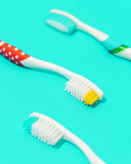 Three toothbrushes are placed on a bright turquoise background. The toothbrushes have white handles with different colored accents. One has red polka dots, another has blue and green stripes, and the last one has a simple cyan accent. The bristles of the brushes are white with some yellow and cyan highlights.