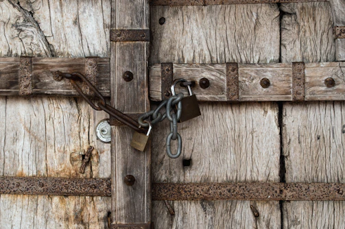 A rustic, weathered wooden door secured with multiple locks and chains. The wood is textured and worn, showing signs of age and exposure to the elements. Metal hinges and fasteners with a rusty appearance are fixed on the door.