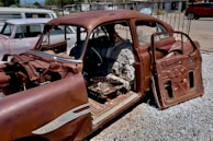 A rusted, partially disassembled vintage car, with its doors open and significant components like the engine and seats missing. The car is parked on gravel, in front of an older vehicle and various buildings in the background.