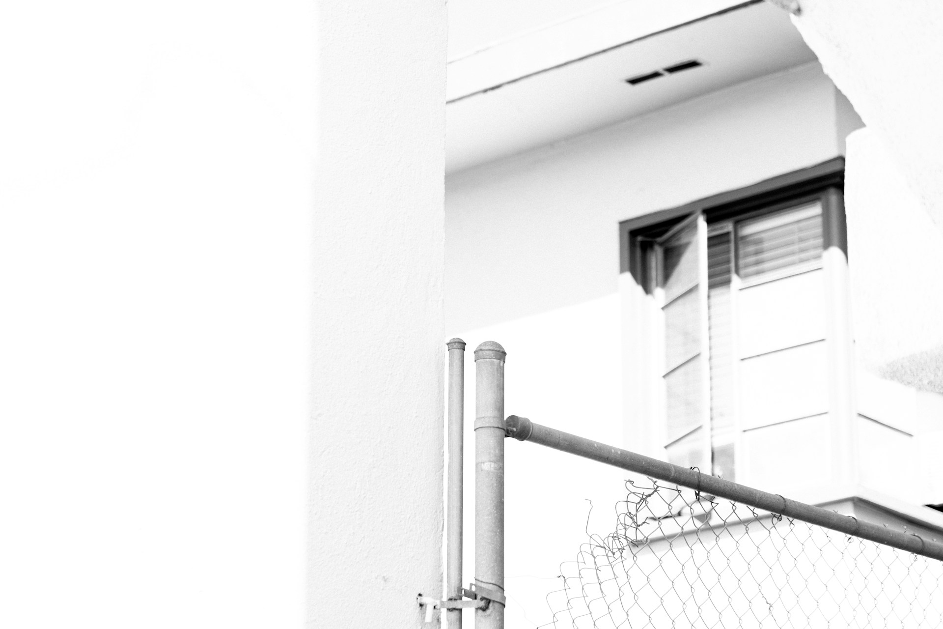 An exterior portion of a building with a partially open window. A chain-link fence is also visible in the foreground. The image is in black and white, with strong contrasts between light and shadow.