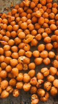 A close-up view of a pile of roasted chickpeas on a flat surface. The chickpeas are golden-brown in color and appear to be seasoned with various spices. They have a crispy texture and some are slightly broken.