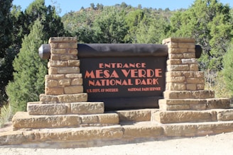 A large wooden sign with stone pillars on each side reads 'Entrance Mesa Verde National Park' with additional text 'U.S. Dept. of Interior National Park Service.' The sign is surrounded by lush green trees and shrubs, suggesting a natural and scenic environment.