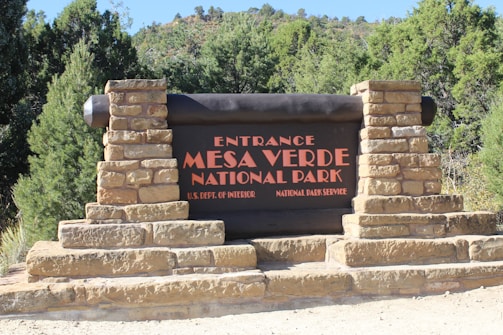 A large wooden sign with stone pillars on each side reads 'Entrance Mesa Verde National Park' with additional text 'U.S. Dept. of Interior National Park Service.' The sign is surrounded by lush green trees and shrubs, suggesting a natural and scenic environment.