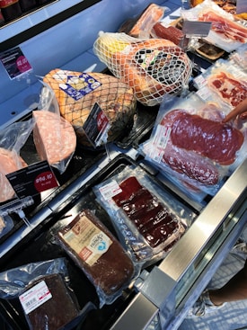 A variety of packaged meats are displayed in a deli section, including ham encased in netting, vacuum-sealed salami, and other cured meats. Price tags are visible, indicating different costs for each product. A hand is holding one packaged item, suggesting selection or purchase.