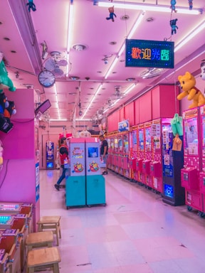 A vibrant arcade with pink and purple lighting fills the space, lined with various claw machines and arcade games. Hanging toys and plushies add to the amusement park-like atmosphere. A couple of people are playing games, adding a lively element to the scene.