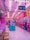 A vibrant arcade with pink and purple lighting fills the space, lined with various claw machines and arcade games. Hanging toys and plushies add to the amusement park-like atmosphere. A couple of people are playing games, adding a lively element to the scene.