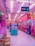 A vibrant arcade with pink and purple lighting fills the space, lined with various claw machines and arcade games. Hanging toys and plushies add to the amusement park-like atmosphere. A couple of people are playing games, adding a lively element to the scene.
