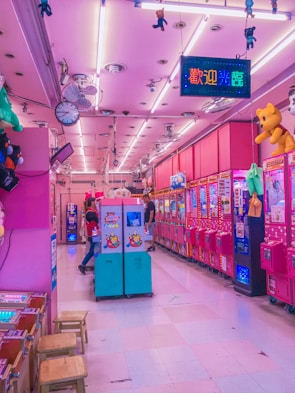 A vibrant arcade with pink and purple lighting fills the space, lined with various claw machines and arcade games. Hanging toys and plushies add to the amusement park-like atmosphere. A couple of people are playing games, adding a lively element to the scene.