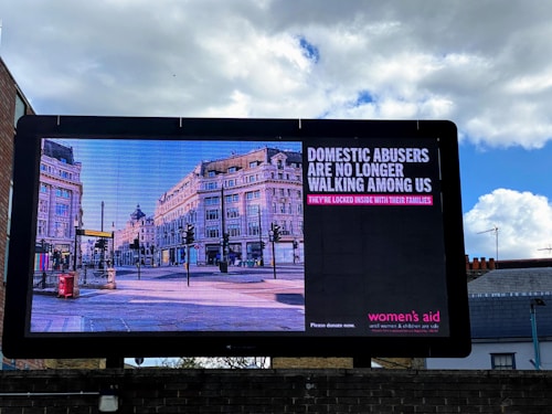 A large billboard displays a public service announcement addressing domestic abuse, featuring an image of a city street lined with historic buildings. The text on the billboard reads: 'Domestic abusers are no longer walking among us. They’re locked inside with their families.'