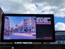A large billboard displays a public service announcement addressing domestic abuse, featuring an image of a city street lined with historic buildings. The text on the billboard reads: 'Domestic abusers are no longer walking among us. They’re locked inside with their families.'