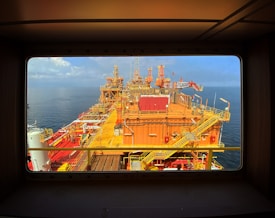 A large offshore oil platform is seen framed by the window of a structure, showcasing various pipes, cranes, and walkways. The platform is orange and surrounded by calm blue ocean under a clear sky.