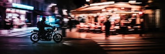 A nighttime street scene featuring a motorcyclist riding past a bustling outdoor caf&eacute;. The image captures the motion of the motorcycle, with blurred streetlights and neon signs creating a sense of speed and energy. Warm lights from the caf&eacute; illuminate patrons seated under a canopy, adding to the vibrant atmosphere.
