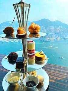 A tiered stand showcasing a variety of elegant pastries and desserts, positioned on a reflective table against a panoramic view of a harbor and cityscape. The upper tiers hold small sandwiches and pastries, while the lower tiers display cakes, a parfait, and small bowls of cream and jam. The background reveals serene blue waters dotted with boats and a city horizon.