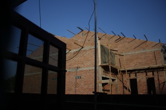 A partially constructed brick building with exposed beams and scaffolding, set against a clear blue sky. The facade is primarily composed of red bricks, with some wooden structures indicating ongoing construction. In the foreground, a window frame partially obstructs the view.