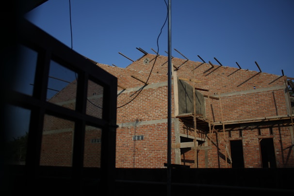 A partially constructed brick building with exposed beams and scaffolding, set against a clear blue sky. The facade is primarily composed of red bricks, with some wooden structures indicating ongoing construction. In the foreground, a window frame partially obstructs the view.