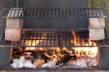A makeshift outdoor barbecue grill setup with flames rising from burning logs and charcoal. Two grill racks are stacked, separated by bricks on each side, allowing for different cooking levels. The fire provides a rustic and warm ambiance.
