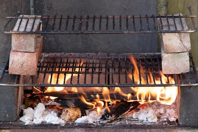 A makeshift outdoor barbecue grill setup with flames rising from burning logs and charcoal. Two grill racks are stacked, separated by bricks on each side, allowing for different cooking levels. The fire provides a rustic and warm ambiance.