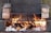 A makeshift outdoor barbecue grill setup with flames rising from burning logs and charcoal. Two grill racks are stacked, separated by bricks on each side, allowing for different cooking levels. The fire provides a rustic and warm ambiance.