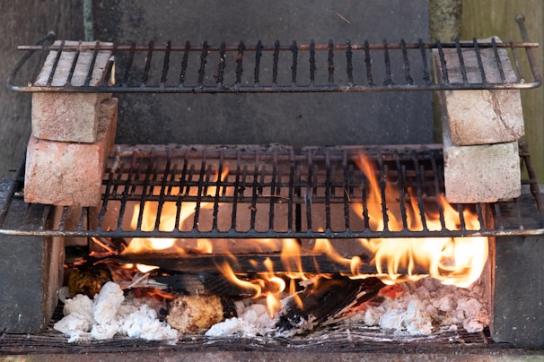 A makeshift outdoor barbecue grill setup with flames rising from burning logs and charcoal. Two grill racks are stacked, separated by bricks on each side, allowing for different cooking levels. The fire provides a rustic and warm ambiance.