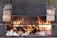 A makeshift outdoor barbecue grill setup with flames rising from burning logs and charcoal. Two grill racks are stacked, separated by bricks on each side, allowing for different cooking levels. The fire provides a rustic and warm ambiance.
