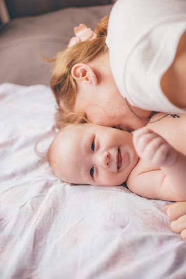 A mother lovingly embraces her baby on a soft white blanket. The baby is smiling and appears content and happy. The scene conveys a sense of warmth and tenderness.