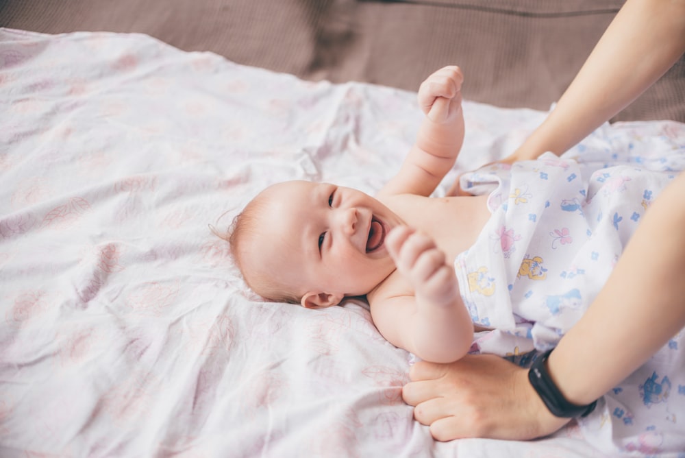 a smiling baby laying on a bed with a woman's arm