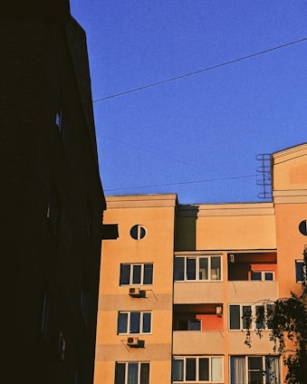 A multi-story residential building with a beige facade is bathed in warm sunlight against a clear blue sky. Several air conditioning units are attached below the windows, which are interspersed across the building. The structure has rectangular and circular architectural elements. In the foreground, part of another building creates a contrasting shadow.