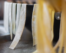 Thin strips of homemade pasta hang over a dark surface, showcasing the texture and slight dusting of flour. The focus highlights the front strands with a gradual blur towards the background.