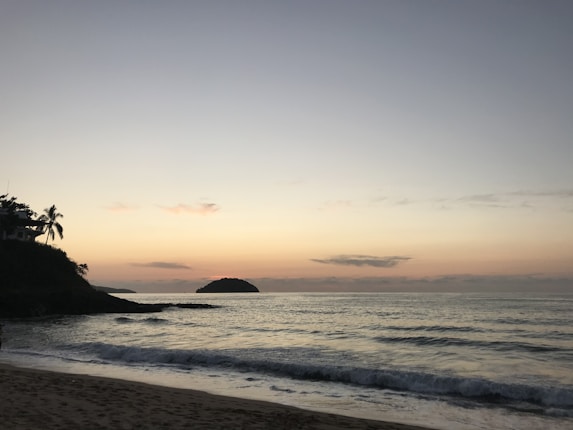 A serene beach scene during sunset with gentle waves lapping against the shore. The sky is a gradient of pastel hues from pale blue to soft orange. A small island is visible in the distance, and the silhouette of palm trees and a structure is on the left side.