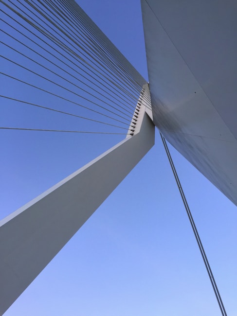 An abstract perspective of a modern bridge with strong geometric lines and cables against a clear blue sky. The image highlights the architectural design and engineering of the structure, emphasizing the tension and symmetry of its components.