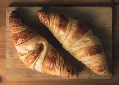 Two golden-brown croissants placed on a wooden cutting board. The flaky pastry has a crispy texture with a glossy finish, indicating a fresh, baked quality.