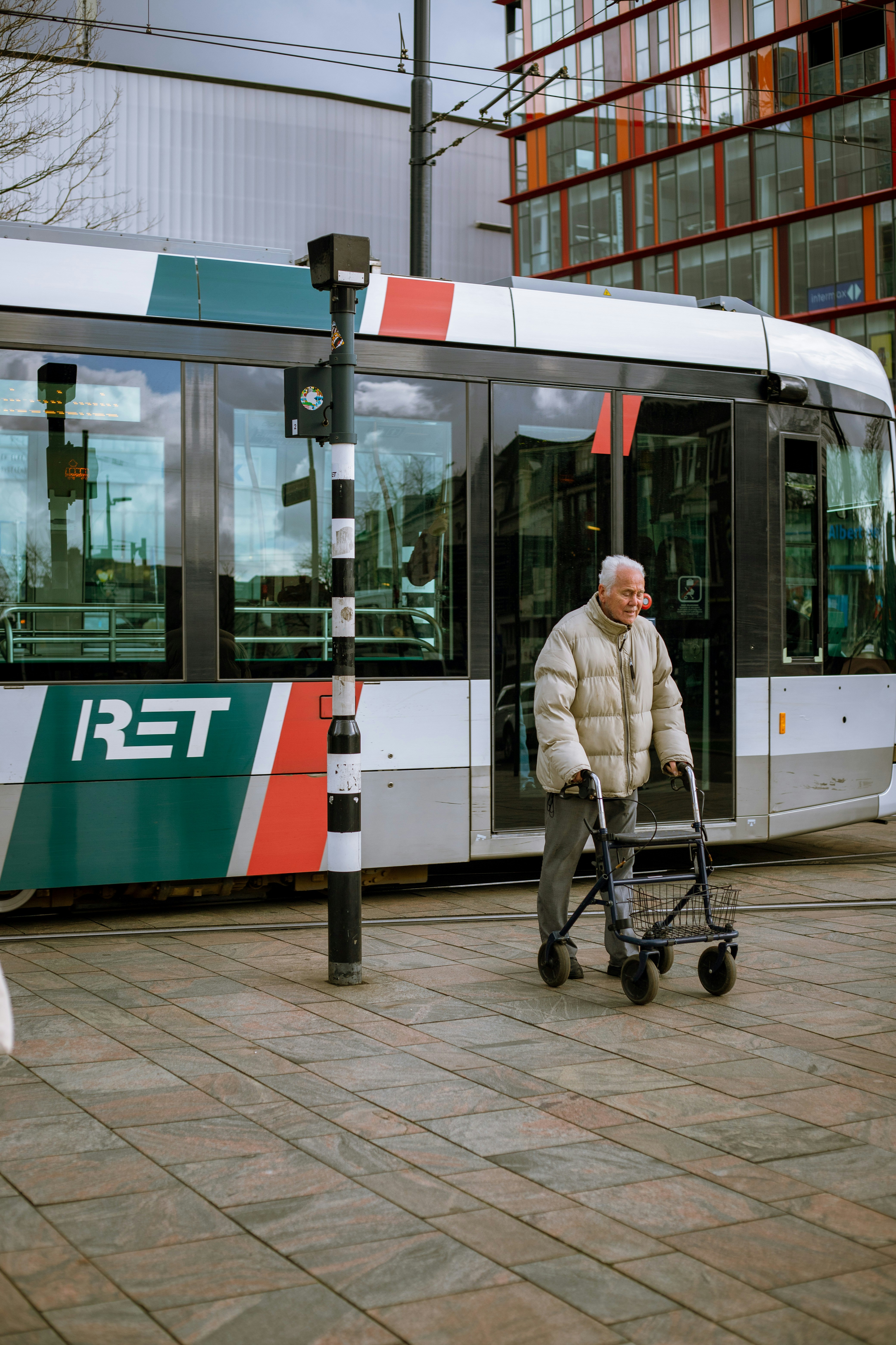 An elderly person using a walker crosses in front of a modern tram with the text 'RET' on its side. The tram is adorned with multiple colors including red, green, and white. In the background, there is a modern building with large glass windows.
