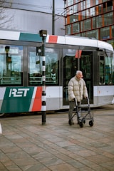 An elderly person using a walker crosses in front of a modern tram with the text 'RET' on its side. The tram is adorned with multiple colors including red, green, and white. In the background, there is a modern building with large glass windows.